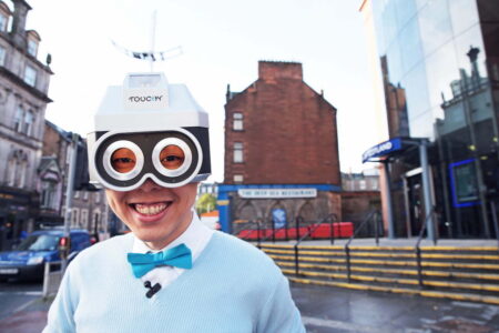 Eric Sui dressed as his alter ego Touchy. He is wearing a white shirt with a blue bow tie. On his head is a silver robot type helmet with black goggles over his eyes. he is standing in front of the Bank of Scotland building in Dundee.