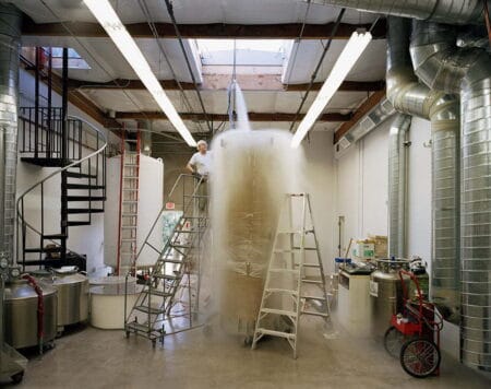 A large workshop. In the centre are two sets of step ladders and a large cylindrical object with what looks like steam coming out from the top of it.