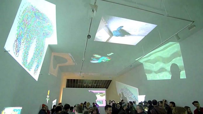 A large audience in a large, white exhibition space. All over the walls and ceiling are various projected images.
