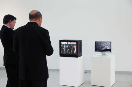 Two people looking at art works in a white exhibition space which contains two computer monitors, each sitting on a white plinth. The monitor on the left has a brightly coloured pattern on the screen, the smaller monitor on the right has white writing on the screen.