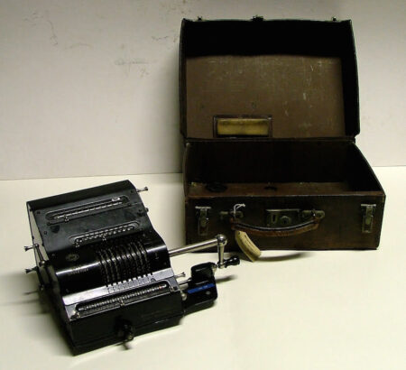 The Brunsviga Nova calculator, which consists of a small metal box containing various dials, switches and levers. Next to this contraption is an empty brown leather case.