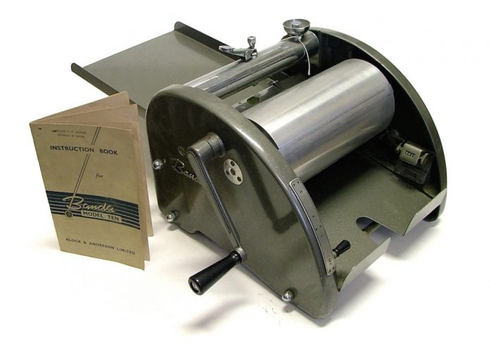 The Spirit Duplicator (or Banda Machine). A semi circle shaped box made of metal, with a handle on the outside which is connected to a cylinder in the centre.