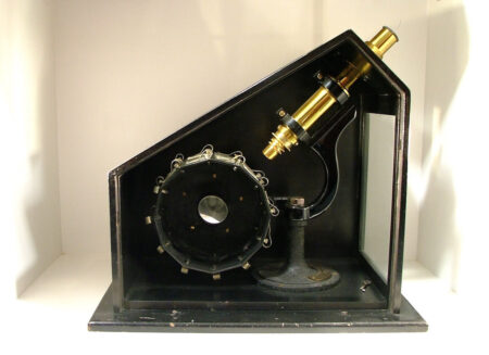 Waterhouse Museum Microscope (for viewing multiple slides). Side view of an old style microscope which consists of a black, metal frame surrounding a black, cylindrical drum which is connected to a brass coloured, cylindrical eye piece.