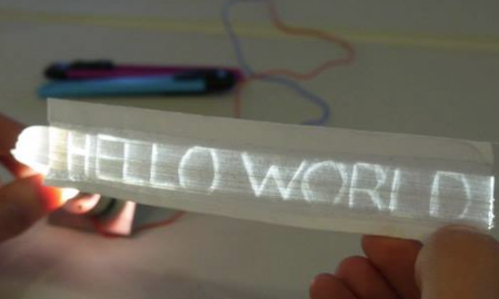A persons fingers holding a small strip of paper with the words " Hello World" illuminated on the surface.