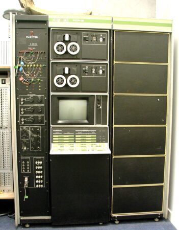 The PDP-12 Programmed Data Processor. A very old style looking computer which consists of a small computer monitor and keyboard surrounded by black boxes, two reel to reel tqpe machines and a series of wires and switches.