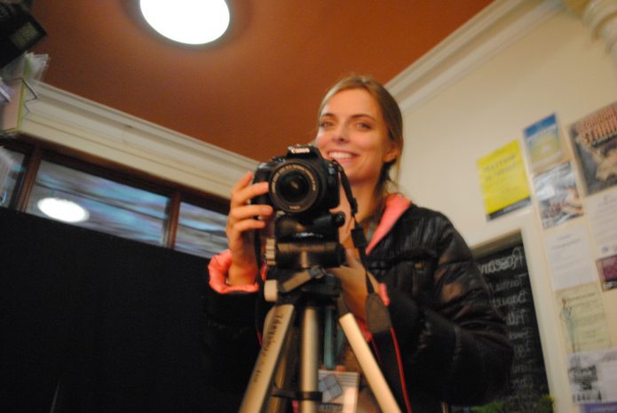 A person smiling whilst standing behind a camera.