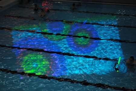 Two coloured circles, one with a blue coloured centre and yellow outer circle, the other circle has a yellow centre with blue outer circle. These circles are projected onto the surface of a swimming pool.