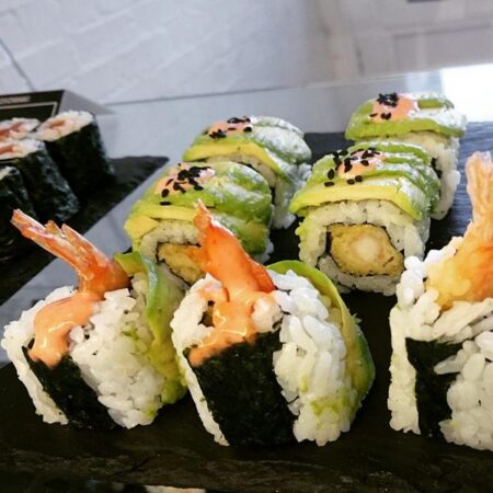Seven pieces of sushi, four of which have are topped with avocado and three have prawns coming out of them.