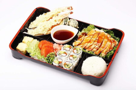 A Japanese Bento box containing sushi, tempura, sliced chicken, rice and a small bowl of soy sauce.