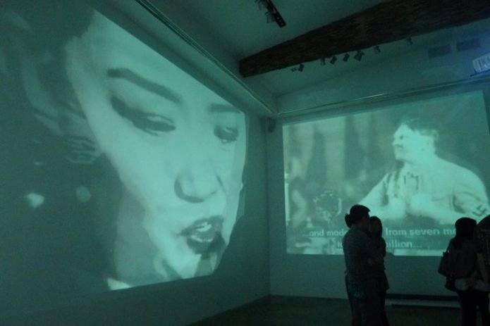 On two walls in an exhibition space are two projected images. The image on the left is of an asian person,possibly Chinese.The image on the right is of Adolf Hitler.