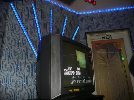 An inside view of a Karaoke room. The walls have silver and blue glittery wallpaper. In the centre of the image is a black, square television with the words " There was no way of knowing" written on the screen.