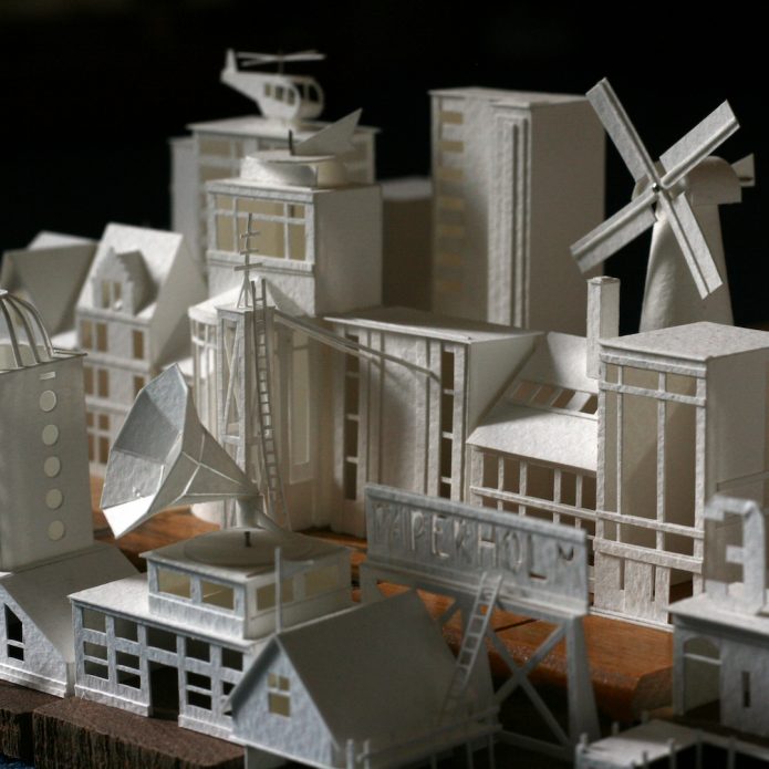A close up shot of a group of model buildings made of paper. These model buildings are white in colour and include various tower blocks, houses and a windmill.