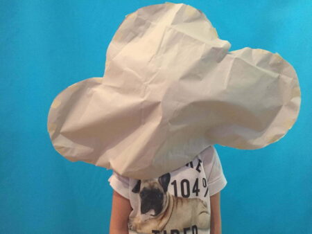 A person standing against a blue background with a large, white cloud shaped object made of paper is placed over their head.