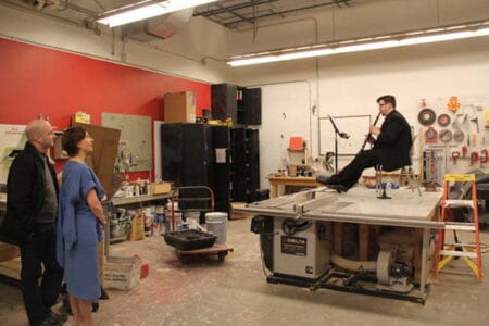 A person playing the clarinet whilst seated on top of a piece of machinery. Two people watch on in one corner of the room.