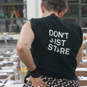 A person standing up at a desk with their back to the camera.The person is wearing a black t-shirt with the words " Don't Just Stare" written in white writing on the back.