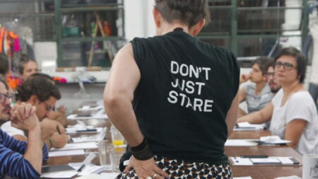 A person standing up at a desk with their back to the camera.The person is wearing a black t-shirt with the words " Don't Just Stare" written in white writing on the back.