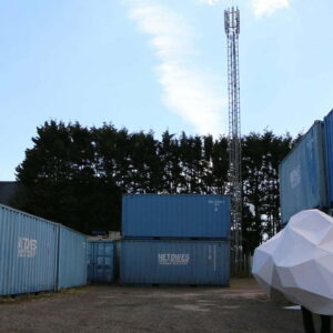 A person wearing a large white shape,made up of various geometric shapes, walks in a yard full of large,blue shipping containers.