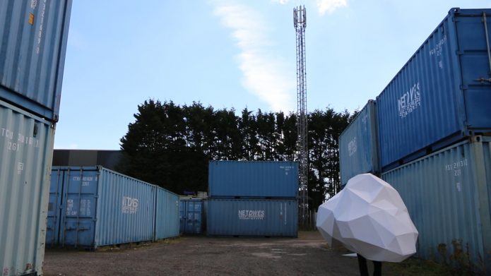 A person wearing a large white shape,made up of various geometric shapes, walks in a yard full of large,blue shipping containers.