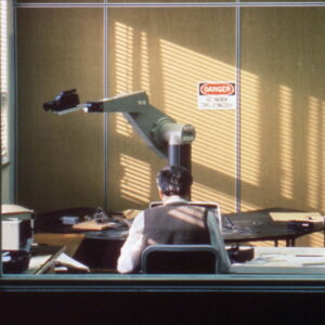 person sits at a desk in an office while a large mechanical arm with a handheld camera watches on.