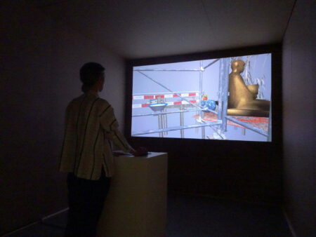 A person in a dark room looking at a large screen. The image on the screen is of a computer generated image of a gold coloured Buddha statue on some scaffolding.