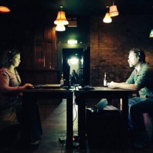 Two people in a darkened room, sitting at a table. One person at one end,the other person at the other end of the table. Each person is wearing headphones and looking at a screen placed at the centre of the table.