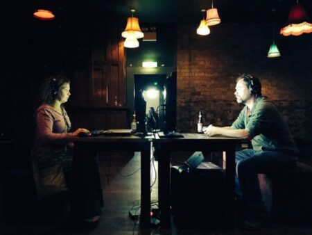 Two people in a darkened room, sitting at a table. One person at one end,the other person at the other end of the table. Each person is wearing headphones and looking at a screen placed at the centre of the table.