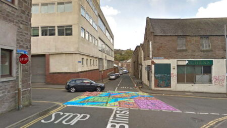 A view of a crossroads in an old industrial estate. Superimposed over the middle of the road is a colourful,computer generated street map.