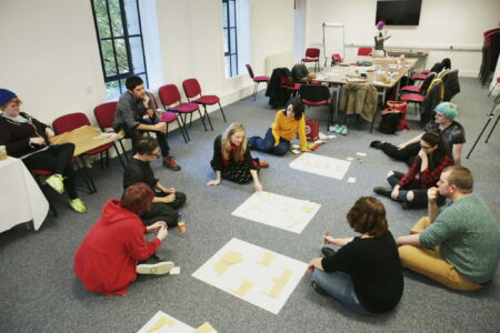 A small group of people,sitting on the floor in a large office space, looking at pieces of paper on the floor.