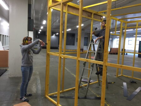Two NEoN volunteers building a large wooden frame in a large empty warehouse.
