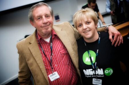 A person wearing a red shirt and a beige jacket putting their arm round another person who is wearing a black and green NEoN t shirt.
