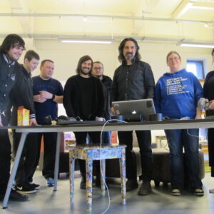 A group of eight people standing behind a table in a brightly lit room, looking at a laptop.