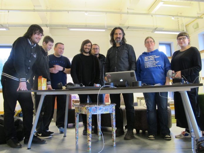 A group of eight people standing behind a table in a brightly lit room, looking at a laptop.
