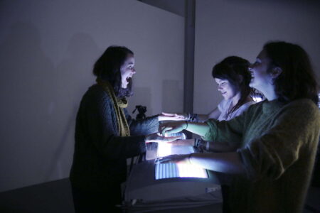 Three people in a dark room holding their hands over brightly lit monitor screens.