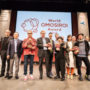 A small group of people, including Japanese sound artists Ei Wada and Usaginingen,standing on a stage in front of a white screen with the words " World Omisiroi Award" written on it.