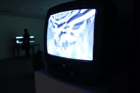 In a dark room a small television monitor sits on a plinth, on the screen is a black and white abstract image.