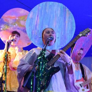Two people playing electric guitar whilst another person sings into a microphone. Each person is wearing a brightly coloured smock and a large circular head piece with a piece cut out for the face.