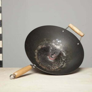 A Wok type frying pan with a plastic bottle glued neck down in the centre, sits on a wooden desk next to a black and white striped stick which is standing vertically.