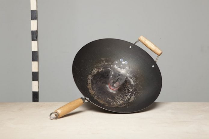 A Wok type frying pan with a plastic bottle glued neck down in the centre, sits on a wooden desk next to a black and white striped stick which is standing vertically.
