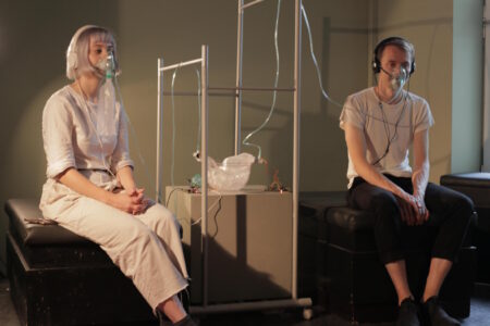 Two people,each sitting on a hospital bed and both wearing oxygen breathing masks which are connected to a bowl situated in the middle of the room.