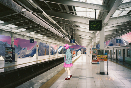 A photographic image of a train station platform. Standing on the platform is an animated image of a person with pink hair,wearing a blue jacket and a pink skirt.