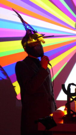 A person wearing a black face mask and a gold headdress with feathers and horns holds a microphone in front of a screen showing a " spectrum" of colours.