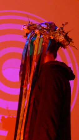 Side view of a person wearing a dark coloured sweatshirt and a headband made of flowers with streamers covering the face. The person is standing in front of a pink and red,illuminated spiral