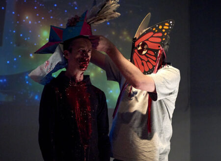 A person wearing a butterfly mask placing a hat made up of geometric shapes,red and blue in colour,onto another persons head.