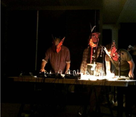Three people,known as Plastique Fantastique, are in a dark room,wearing butterfly masks and looking at various electronic instruments on a lamplit table.