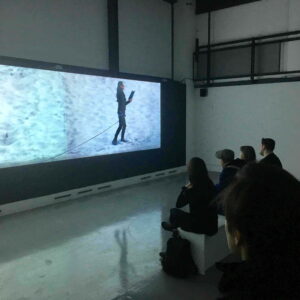 A small audience looking at a long,thin cinema screen. On the screen is a person dressed in black,reading a book and pulling an unseen object with a rope attached to their waist. The background looks like blue and green ice.
