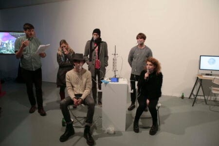 A person reads to five people in a white room whilst they are wearing breathing apparatus thats attached to a small contraption which is situated on a white plinth.