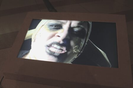 An image of a screen showing a person wearing black lipstick and black and yellow make-up over the eyes.