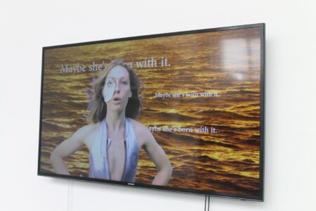 A flat screen television on a white wall. The image on the screen is of a person wearing a silver top and over their right eye is a piece of paper with an eye printed on it. In the background is a sunlit sea and the words " Maybe she's born with it" written over the top in white writing.