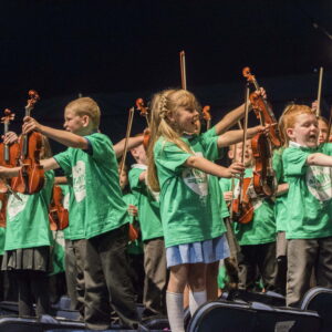 A group of school children,wearing green t-shirts, holding violins and violin bows in the air.