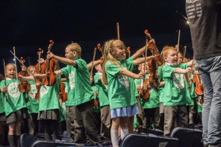A group of school children,wearing green t-shirts, holding violins and violin bows in the air.